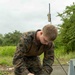 A military service member in MARPAT (Marine pattern) camouflage uniform is working on what appears to be a small fixed-wing UAV (unmanned aerial vehicle), likely a RQ-11 Raven or similar hand-launched drone. The individual is assembling or inspecting the aircraft's wing components on top of a hard-shell Pelican-style transport case. The setting appears to be an outdoor field environment with lush green vegetation in the background.
