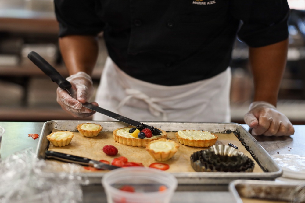 A food service worker wearing a black uniform with 'Marine Aide' text, a white apron, and clear plastic gloves is decorating fruit tarts on a metal baking sheet lined with parchment paper. The worker is using tongs or a long utensil to place mixed berries (blueberries, raspberries) and fruit pieces onto cream-filled pastry tarts. The setting appears to be a professional kitchen environment.