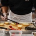A food service worker wearing a black uniform with 'Marine Aide' text, a white apron, and clear plastic gloves is decorating fruit tarts on a metal baking sheet lined with parchment paper. The worker is using tongs or a long utensil to place mixed berries (blueberries, raspberries) and fruit pieces onto cream-filled pastry tarts. The setting appears to be a professional kitchen environment.