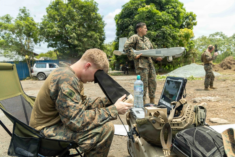 Three military personnel in MARPAT (Marine pattern) camouflage uniforms are conducting what appears to be an unmanned aerial vehicle (UAV) operation in an outdoor setting. One soldier is seated operating a ruggedized laptop control station with a sun shield, a second is holding a fixed-wing drone (likely an RQ-20 Puma or similar small UAV), and a third is standing in the background holding a handheld device. The environment appears to be a semi-rural or forward operating area with trees and a vehicle visible in the background.