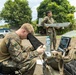 Three military personnel in MARPAT (Marine pattern) camouflage uniforms are conducting what appears to be an unmanned aerial vehicle (UAV) operation in an outdoor setting. One soldier is seated operating a ruggedized laptop control station with a sun shield, a second is holding a fixed-wing drone (likely an RQ-20 Puma or similar small UAV), and a third is standing in the background holding a handheld device. The environment appears to be a semi-rural or forward operating area with trees and a vehicle visible in the background.