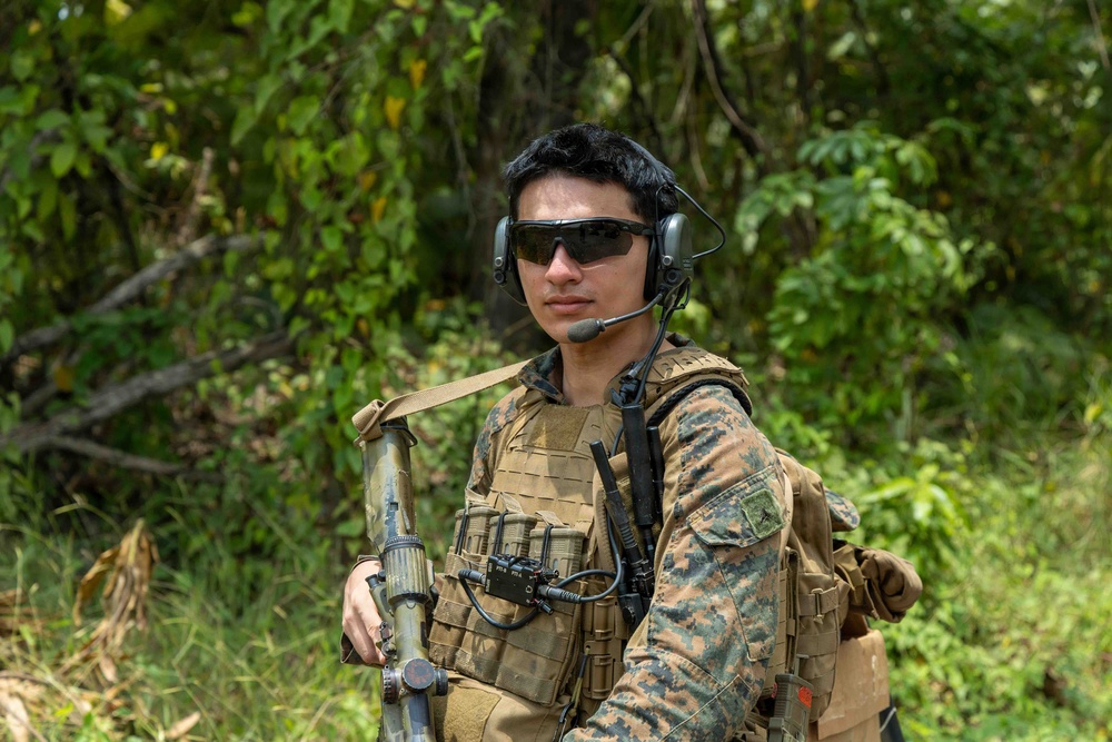 A military service member in MARPAT (Marine pattern) digital camouflage uniform is photographed in a jungle or forested environment. The individual is equipped with tactical gear including a plate carrier vest, communications headset with boom microphone, ballistic sunglasses, and is carrying what appears to be a rifle with a sling. The setting and uniform pattern suggest a U.S. Marine Corps field operation or training exercise.