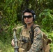A military service member in MARPAT (Marine pattern) digital camouflage uniform is photographed in a jungle or forested environment. The individual is equipped with tactical gear including a plate carrier vest, communications headset with boom microphone, ballistic sunglasses, and is carrying what appears to be a rifle with a sling. The setting and uniform pattern suggest a U.S. Marine Corps field operation or training exercise.
