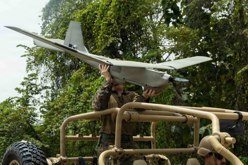 A military service member in camouflage uniform is handling a fixed-wing unmanned aerial vehicle (UAV/drone) while standing on or beside a tan/desert-colored military vehicle. The setting appears to be a tropical or jungle environment based on the dense green foliage in the background. The service member appears to be preparing the UAV for launch or recovering it after a mission.