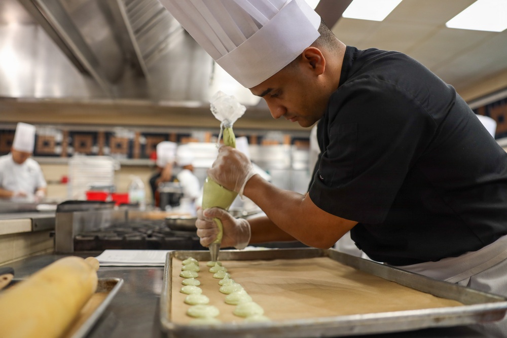 A chef in a black uniform and white toque is carefully piping a green-colored filling or batter onto a parchment-lined baking sheet in a professional kitchen. The foreground subject is in sharp focus while other kitchen staff are visible but blurred in the background. The setting appears to be a culinary school or high-volume professional kitchen.