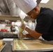 A chef in a black uniform and white toque is carefully piping a green-colored filling or batter onto a parchment-lined baking sheet in a professional kitchen. The foreground subject is in sharp focus while other kitchen staff are visible but blurred in the background. The setting appears to be a culinary school or high-volume professional kitchen.