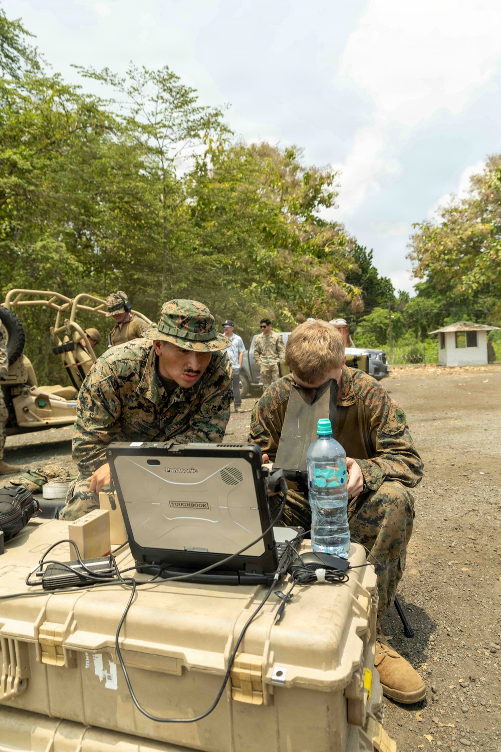 Two military personnel in MARPAT digital camouflage uniforms are working together on a Panasonic Toughbook rugged laptop placed on a tan Pelican-style hard case. The setting appears to be an outdoor field environment with military vehicles, additional personnel, and tropical vegetation visible in the background. The scene suggests a field operations or training exercise involving tactical computing equipment.