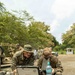 Two military personnel in MARPAT digital camouflage uniforms are working together on a Panasonic Toughbook rugged laptop placed on a tan Pelican-style hard case. The setting appears to be an outdoor field environment with military vehicles, additional personnel, and tropical vegetation visible in the background. The scene suggests a field operations or training exercise involving tactical computing equipment.