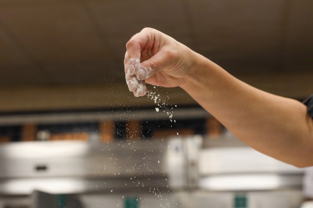 A hand is captured mid-action sprinkling or tossing a white powdery substance, likely flour or salt, over a stainless steel surface in what appears to be a commercial kitchen. The motion is frozen in time, showing particles of the white substance falling downward. The background contains blurred kitchen equipment consistent with a professional cooking environment.