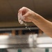 A hand is captured mid-action sprinkling or tossing a white powdery substance, likely flour or salt, over a stainless steel surface in what appears to be a commercial kitchen. The motion is frozen in time, showing particles of the white substance falling downward. The background contains blurred kitchen equipment consistent with a professional cooking environment.