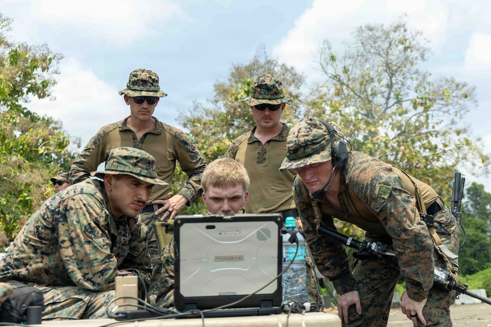 A group of military personnel in camouflage uniforms are gathered around a ruggedized laptop computer, appearing to review data or coordinate operations. The setting appears to be an outdoor field environment with trees visible in the background. The personnel are wearing U.S. Marine Corps MARPAT digital camouflage uniforms and tactical gear.