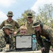 A group of military personnel in camouflage uniforms are gathered around a ruggedized laptop computer, appearing to review data or coordinate operations. The setting appears to be an outdoor field environment with trees visible in the background. The personnel are wearing U.S. Marine Corps MARPAT digital camouflage uniforms and tactical gear.