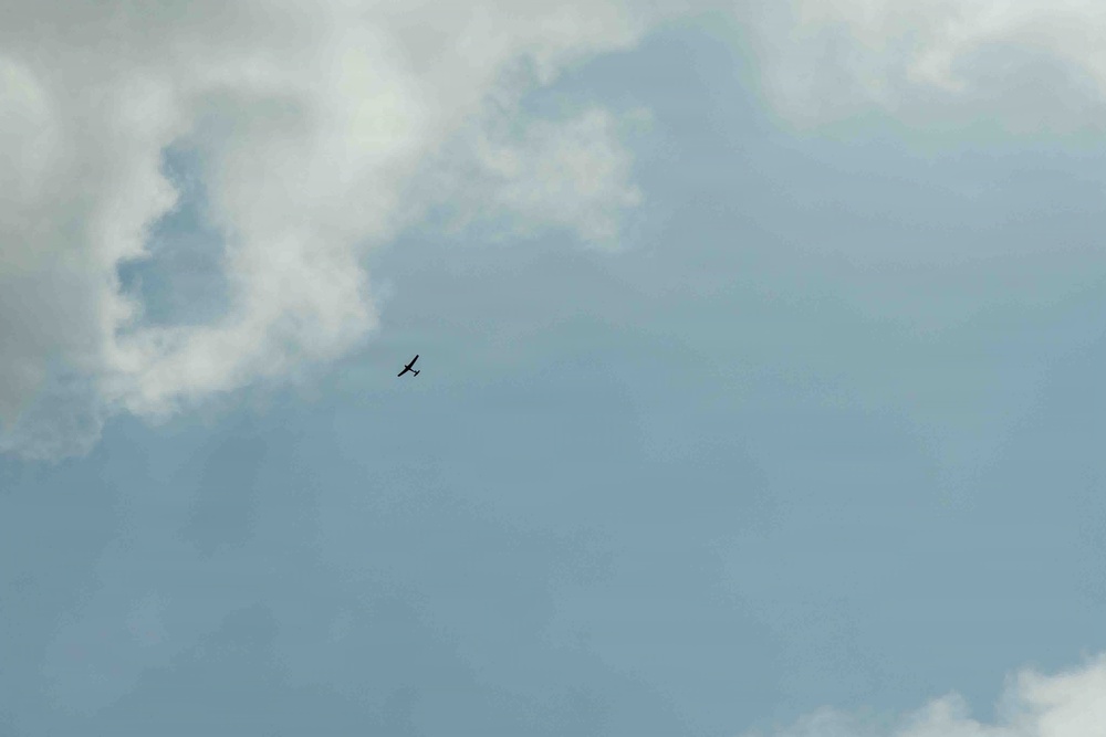 The image shows a wide sky scene dominated by blue sky and scattered white-grey clouds. A single small dark silhouette of what appears to be a small aircraft or large bird is visible in the upper-center-left portion of the frame. The subject is very small relative to the expansive sky background.