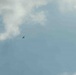 The image shows a wide sky scene dominated by blue sky and scattered white-grey clouds. A single small dark silhouette of what appears to be a small aircraft or large bird is visible in the upper-center-left portion of the frame. The subject is very small relative to the expansive sky background.
