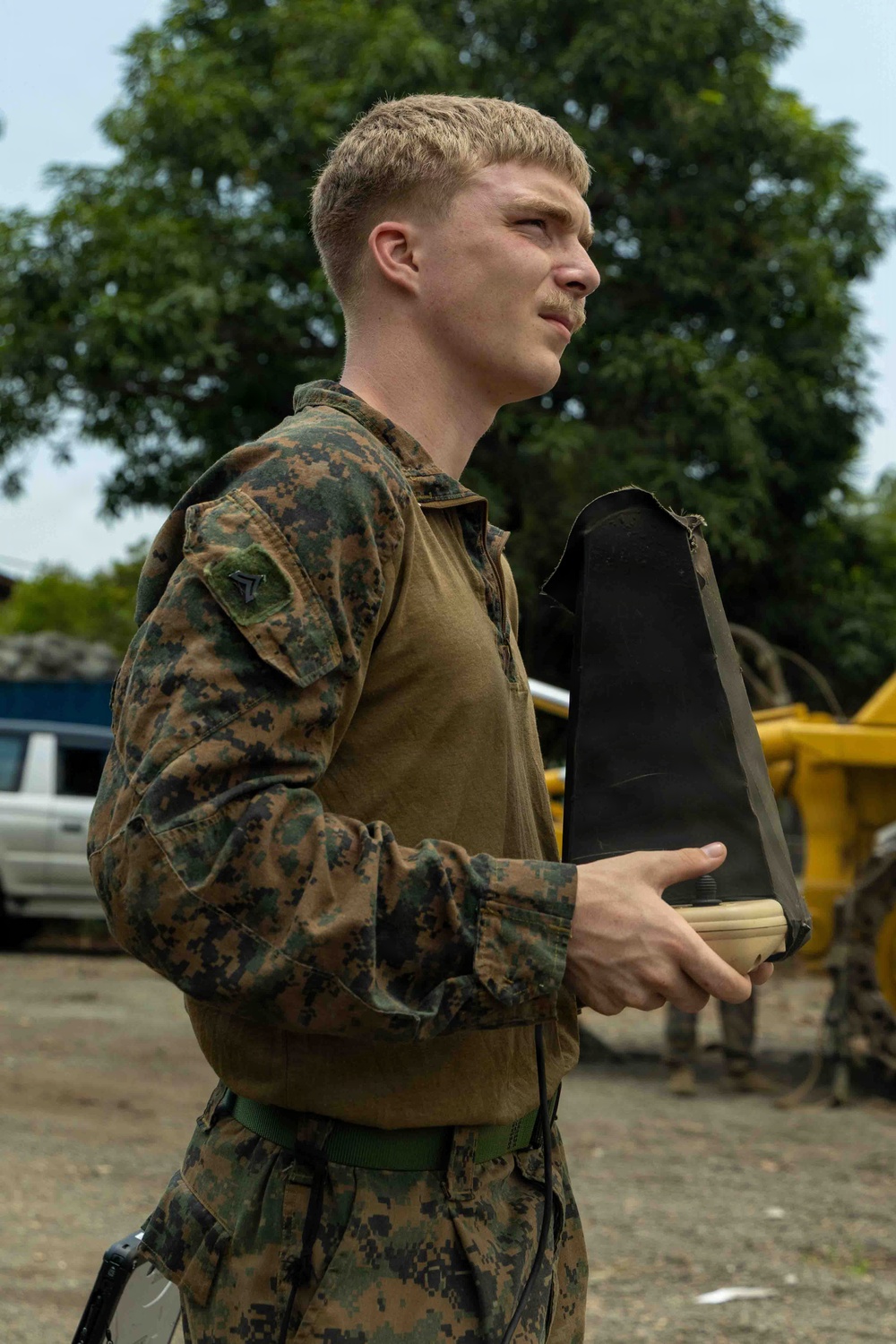 A young male service member in U.S. Marine Corps MARPAT woodland camouflage uniform is photographed in a side profile view. He is holding a tan/black piece of equipment, possibly a drone controller or electronic device with a protective cover. The setting appears to be an outdoor working area with vehicles and heavy equipment visible in the background.