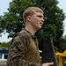 A young male service member in U.S. Marine Corps MARPAT woodland camouflage uniform is photographed in a side profile view. He is holding a tan/black piece of equipment, possibly a drone controller or electronic device with a protective cover. The setting appears to be an outdoor working area with vehicles and heavy equipment visible in the background.