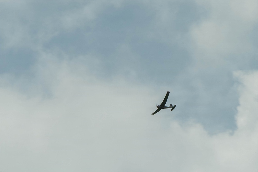 A small aircraft, likely a glider or light plane, is captured in flight against an overcast, cloudy sky. The aircraft appears to be banking or turning, visible in silhouette against the grey-white cloud cover. The image was taken from a ground-level perspective looking upward.