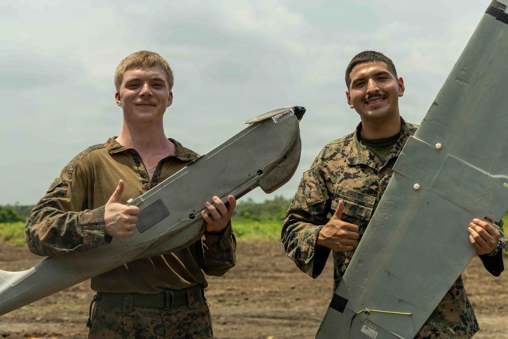 Two U.S. Marines in MARPAT camouflage uniforms are posing with a fixed-wing unmanned aerial vehicle (UAV), both giving thumbs-up gestures. They appear to be on a dirt field or airstrip with vegetation visible in the background. The mood is celebratory, suggesting a successful UAV operation or training exercise.