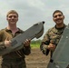 Two U.S. Marines in MARPAT camouflage uniforms are posing with a fixed-wing unmanned aerial vehicle (UAV), both giving thumbs-up gestures. They appear to be on a dirt field or airstrip with vegetation visible in the background. The mood is celebratory, suggesting a successful UAV operation or training exercise.