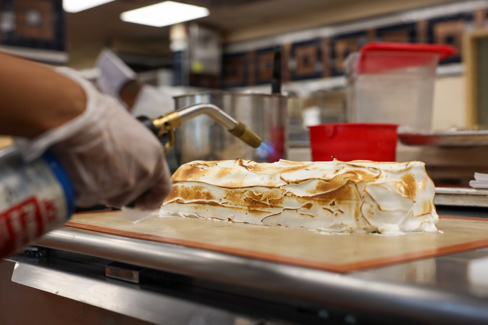 A professional kitchen worker is using a culinary torch to toast the meringue topping on a layered dessert, likely a Baked Alaska or meringue cake. The dessert sits on a silicone baking mat on a stainless steel surface. The background shows a commercial kitchen environment with various containers and equipment.
