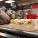 A professional kitchen worker is using a culinary torch to toast the meringue topping on a layered dessert, likely a Baked Alaska or meringue cake. The dessert sits on a silicone baking mat on a stainless steel surface. The background shows a commercial kitchen environment with various containers and equipment.