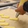 A person is piping choux pastry dough onto a parchment-lined baking sheet using a yellow pastry bag fitted with a metal tip. Several elongated strips of pale yellow dough have already been piped onto the tray, consistent with éclair or ladyfinger preparation. The scene appears to be in a professional or semi-professional kitchen setting.