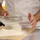 A chef in a white uniform is sifting a white powdery substance, likely flour or powdered sugar, through a fine mesh strainer onto parchment paper placed on a stainless steel work surface. The action captures the powder mid-fall, creating a dynamic visual of the sifting process. The setting appears to be a professional kitchen environment.