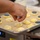 A person is preparing baked goods on a parchment-lined sheet pan in what appears to be a professional kitchen. They are sprinkling shredded cheese (likely Parmesan) onto piped mounds of yellow dough or batter. Some of the dough mounds are topped with dark blue circular discs, possibly portioning guides or flavor markers.