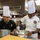 Two United States Marine Corps personnel wearing chef's toques and culinary uniforms are working together in a professional kitchen. The individual on the left wears a black Marine Corps uniform with sergeant chevrons, while the one on the right wears a white chef's coat with a USMC emblem patch. They appear to be preparing food in mixing bowls in what looks like a large institutional or military kitchen.