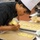 A pastry chef wearing a white toque and black chef's jacket is carefully piping a yellow batter or dough onto a parchment-lined baking sheet. The chef is leaning in closely to ensure precision while working in a professional kitchen environment. A second baking sheet in the background already contains rows of piped dough pieces ready for baking.
