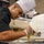 A young male chef wearing a white toque and black chef's jacket is focused on piping a yellow filling or batter from a large plastic piping bag onto a baking tray. He is wearing disposable gloves and working in what appears to be a professional or institutional kitchen. The image captures a moment of precise culinary work with sharp focus on the subject.