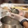 A person wearing disposable plastic gloves is kneading or shaping a dark green dough or paste over a stainless steel mixing bowl. The setting appears to be a professional or semi-professional kitchen environment. The green color of the dough suggests it may contain ingredients such as matcha, spinach, or other green herbs.