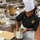 A person wearing a black chef's jacket embroidered with a U.S. Marine Corps emblem and 'Marine Aide' text works with a stainless steel mixing bowl in a professional kitchen. They are wearing a white toque (chef's hat) and a smartwatch, and appear to be preparing or mixing ingredients. Several other kitchen staff in white uniforms are visible working in the background.