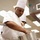 A chef in full white uniform, including a tall toque blanche, is working in a professional kitchen environment. He is focused downward, handling a large stainless steel stockpot. At least one other person in chef's whites is visible in the background.