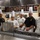 Five culinary professionals wearing chef's toques and uniforms are gathered around a commercial kitchen workstation. They appear to be engaged in a cooking demonstration or training session, with a large stockpot visible on the range. Several individuals are wearing emblems consistent with U.S. Marine Corps insignia on their uniforms.