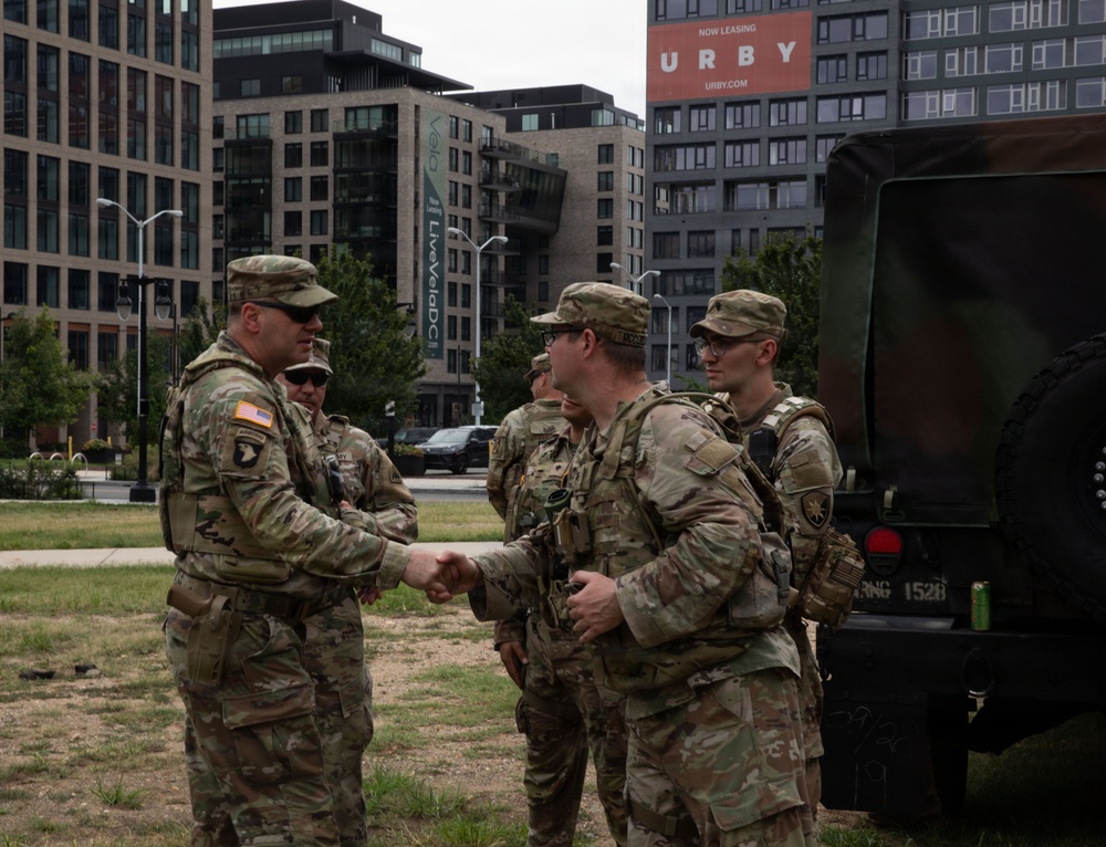 A group of U.S. Army soldiers in OCP (Operational Camouflage Pattern) uniforms are gathered in an urban outdoor area, with one senior soldier shaking hands with another. A military vehicle is visible to the right, and modern urban buildings including a 'URBY' branded structure are visible in the background. The setting appears to be Washington D.C. based on the 'LiveVelaDC' signage visible on one of the buildings.