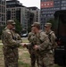 A group of U.S. Army soldiers in OCP (Operational Camouflage Pattern) uniforms are gathered in an urban outdoor area, with one senior soldier shaking hands with another. A military vehicle is visible to the right, and modern urban buildings including a 'URBY' branded structure are visible in the background. The setting appears to be Washington D.C. based on the 'LiveVelaDC' signage visible on one of the buildings.
