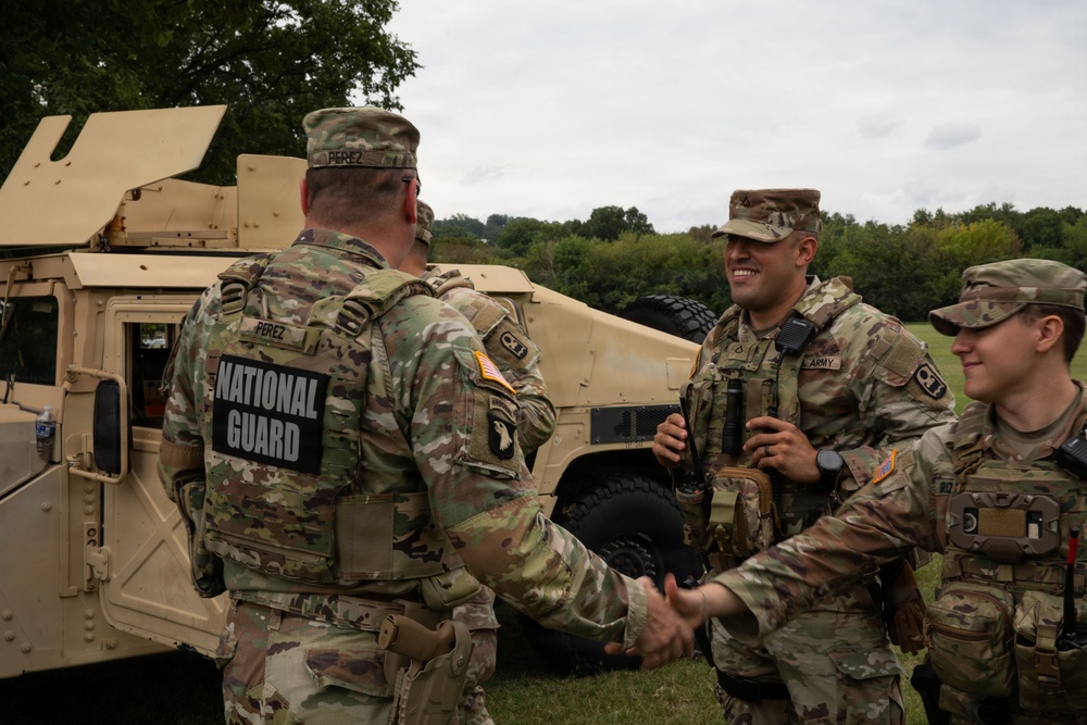 Three U.S. Army National Guard soldiers are gathered near a tan military HMMWV (Humvee) in an outdoor field setting. The soldier facing away from the camera, identified by name tape as 'PEREZ,' is shaking hands with another soldier while a third looks on smiling. The scene appears to be a friendly greeting or recognition moment during a field exercise or operation.