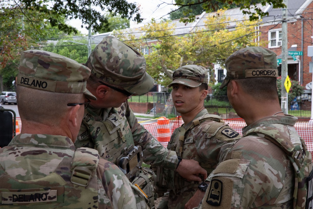 Four U.S. Army soldiers in OCP (Operational Camouflage Pattern) uniforms are gathered in a huddle on a residential street, appearing to be in a briefing or discussion. Name tapes visible include 'DELANO' and 'CORDERO', and at least one soldier has an 'ARMY' tape visible. The setting appears to be an urban neighborhood with brick buildings, trees, and orange construction barriers in the background.