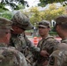 Four U.S. Army soldiers in OCP (Operational Camouflage Pattern) uniforms are gathered in a huddle on a residential street, appearing to be in a briefing or discussion. Name tapes visible include 'DELANO' and 'CORDERO', and at least one soldier has an 'ARMY' tape visible. The setting appears to be an urban neighborhood with brick buildings, trees, and orange construction barriers in the background.