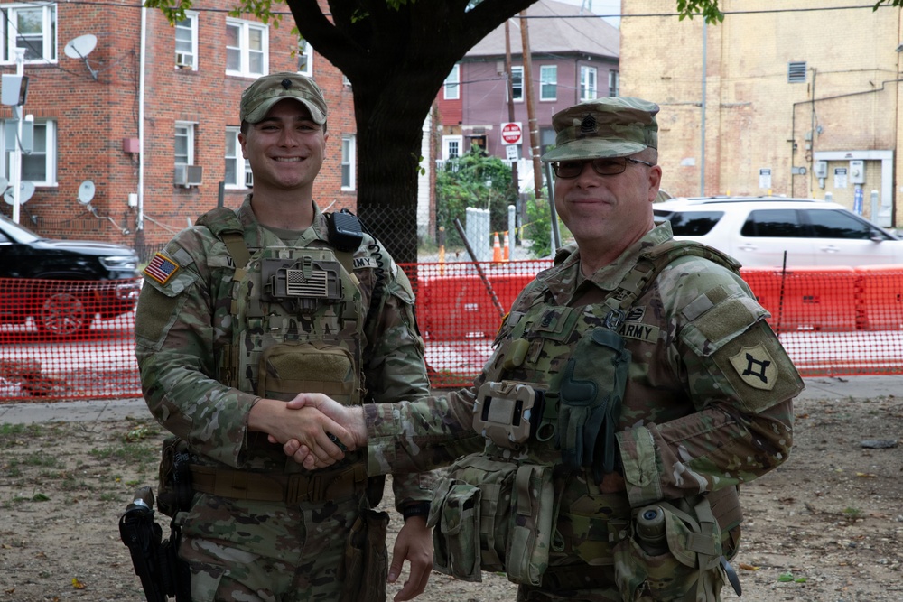 Two U.S. Army soldiers in MultiCam/OCP camouflage uniforms are shaking hands in an urban outdoor setting. Both are wearing tactical vests and patrol caps, with 'ARMY' tape visible on their uniforms. The background shows a residential urban area with brick buildings, orange construction fencing, and parked vehicles.