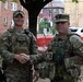 Two U.S. Army soldiers in MultiCam/OCP camouflage uniforms are shaking hands in an urban outdoor setting. Both are wearing tactical vests and patrol caps, with 'ARMY' tape visible on their uniforms. The background shows a residential urban area with brick buildings, orange construction fencing, and parked vehicles.