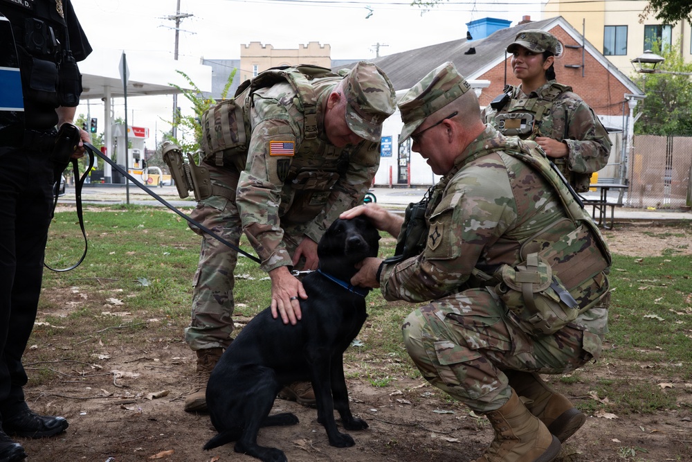 Two U.S. Army soldiers in OCP (Operational Camouflage Pattern) uniforms crouch down to interact with a black Labrador dog on a leash in an urban park setting. A third soldier stands in the background observing, while a law enforcement officer in black tactical gear is partially visible on the left. The scene appears to be a community engagement or patrol operation in a residential neighborhood.