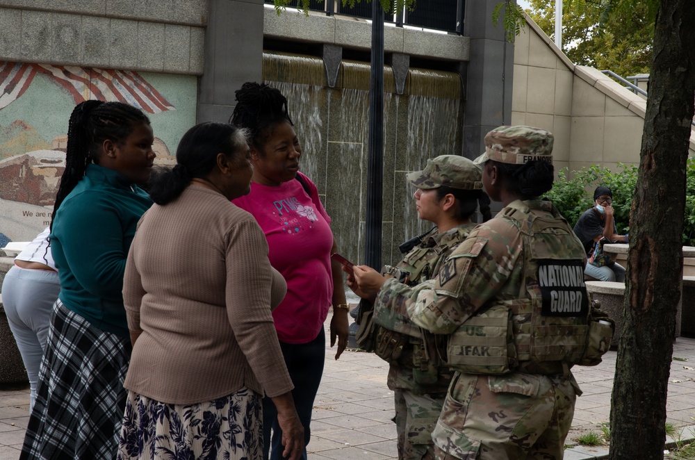 Two National Guard soldiers in camouflage uniforms are interacting with three civilian women in an outdoor urban plaza setting. One soldier's name tape reads 'FRANCOIS' and their vest is labeled 'NATIONAL GUARD.' The scene appears to be a community engagement or information-sharing interaction near a building with a decorative mural and water feature.