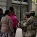 Two National Guard soldiers in camouflage uniforms are interacting with three civilian women in an outdoor urban plaza setting. One soldier's name tape reads 'FRANCOIS' and their vest is labeled 'NATIONAL GUARD.' The scene appears to be a community engagement or information-sharing interaction near a building with a decorative mural and water feature.