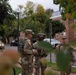 Three U.S. Army National Guard soldiers in OCP (Operational Camouflage Pattern) uniforms are gathered on a sidewalk in what appears to be a residential or mixed-use urban area. The soldiers appear to be in conversation, possibly conducting a briefing or coordination. The image is taken through tree foliage, partially obscuring the view.
