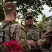 Three National Guard soldiers in OCP (Operational Camouflage Pattern) uniforms are engaged in a conversation outdoors beneath a tree. The soldier facing away from the camera has the name tape 'GREER' and a 'NATIONAL GUARD' patch visible on their back. The setting appears to be a casual outdoor briefing or discussion, with flowering plants visible in the foreground.