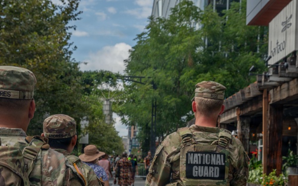 Three U.S. National Guard soldiers in full camouflage uniforms and tactical gear are patrolling a pedestrian walkway lined with trees and commercial establishments. They are walking away from the camera, with name tapes and a 'NATIONAL GUARD' patch clearly visible on their backs. The setting appears to be an outdoor shopping or entertainment district during daytime.