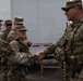A group of U.S. Army soldiers in OCP (Operational Camouflage Pattern) uniforms are engaged in a handshake greeting or recognition ceremony outdoors. The soldier on the right, wearing tactical gear and sunglasses, is shaking hands with another soldier while additional personnel observe. A white specialty trailer with the website 'www.RichSpecialtyTrailers.com' is visible in the background.
