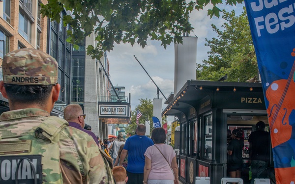 A National Guard soldier with the name tag 'ANDRES' is seen from behind, patrolling a busy outdoor pedestrian area during what appears to be a public festival. The scene includes signage for the DC Jazz Festival and an ABFF (American Black Film Festival) Pop Up event. Civilians, including families with children, are walking along a brick-paved street lined with vendors and shops.